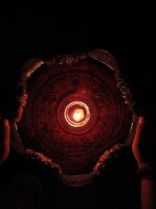 A candle is lit at the center of an ornate lamp being held in the dark, casting a warm glow on the decorative patterns and details of the lamp.