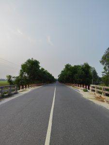 A straight paved road flanked by trees on both sides stretches into the distance under a clear blue sky. Red and white striped markers are visible along the roadside, and a concrete railing lines the edge of a bridge section.