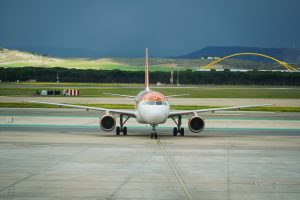 A commercial airplane is positioned and facing the viewer, showcasing a white body with orange accents. The aircraft is situated on a runway with visible markings and green taxiways.
