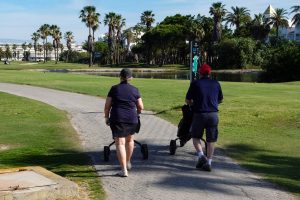 A man and a woman are walking along a paved path in a sunny golf course setting. Each of them pushing a golf cart with wheels. They are both dressed casually. In the background, there are tall palm trees and green grass.