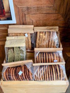 A wooden display bin filled with various packages of freshly baked cookies and biscotti sealed in brown paper bags.