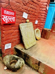 Traditional stone grinding tools displayed next to a bright red brick wall with a sign that reads “Museum Open” in Nepali.