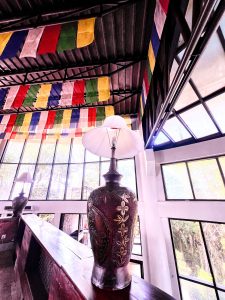 A tall decorative table lamp inside a building with large glass windows and colorful Buddhist prayer flags hanging from the ceiling.