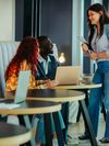 A diverse group of young adults in modern study lounge. They are holding study materials and using laptops; three of the students are engaged in conversation while another young woman works alone.