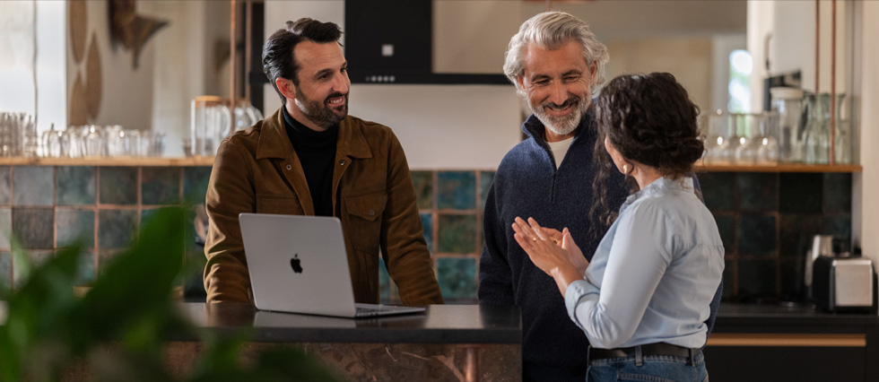 Un MacBook est posé sur une table et deux hommes et une femme discutent ensemble.