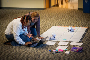 teacher-training-2 Educators working with a laptop on the floor next to a large map where they are guiding robots on a path.