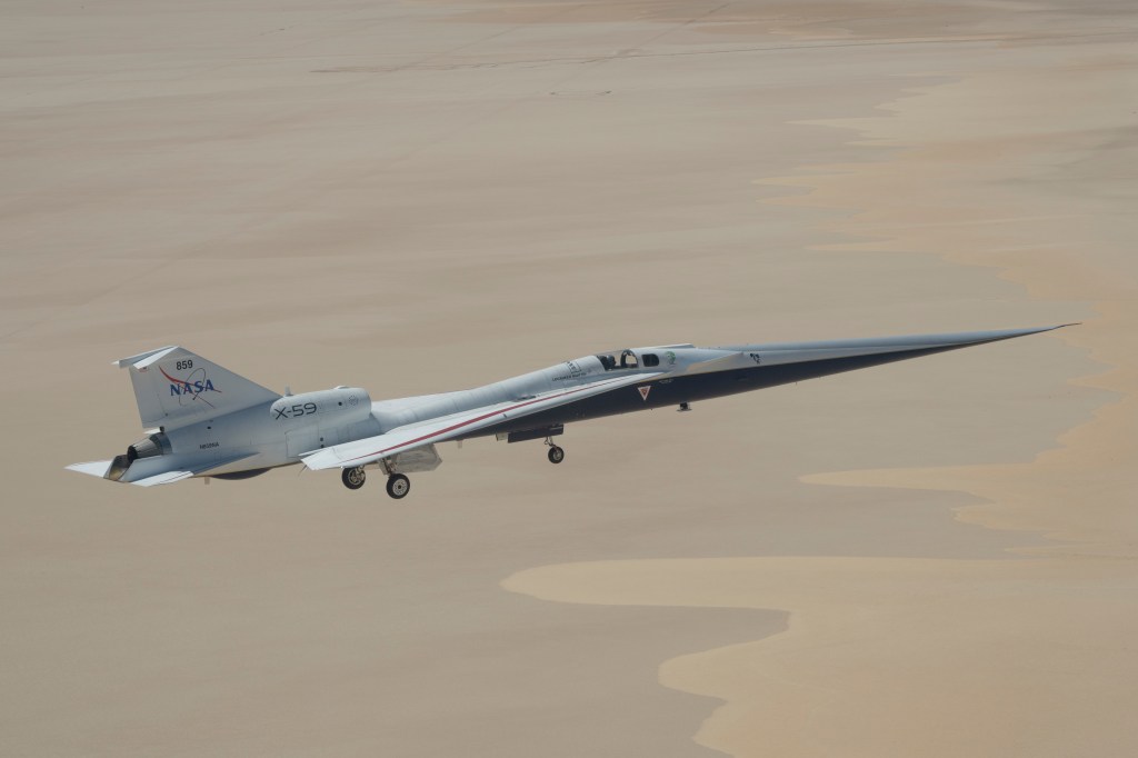 NASA’s X-59 quiet supersonic research aircraft flying above Rogers Dry Lake in California with its landing gear extended during a test flight on May 12, 2026. The aircraft’s long, nose and white fuselage are visible against the light tan desert surface below.
