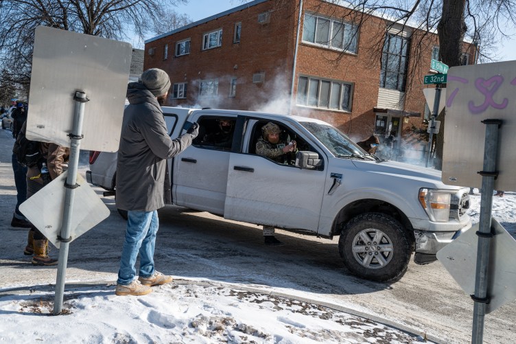 Men in camo wearing helmets and masks open the doors of a four-door pickup truck while holding weapons and a gas canister. Outside the truck, a few people in street clothes are gathered in a snowy suburban roundabout. One is filming on his phone.