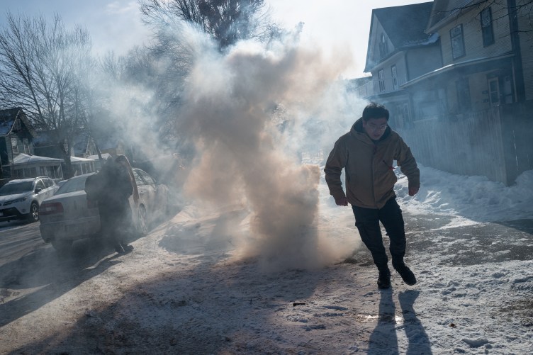 Two people scatter as a column of gas rises from a spot in the middle of a snowy residential sidewalk.