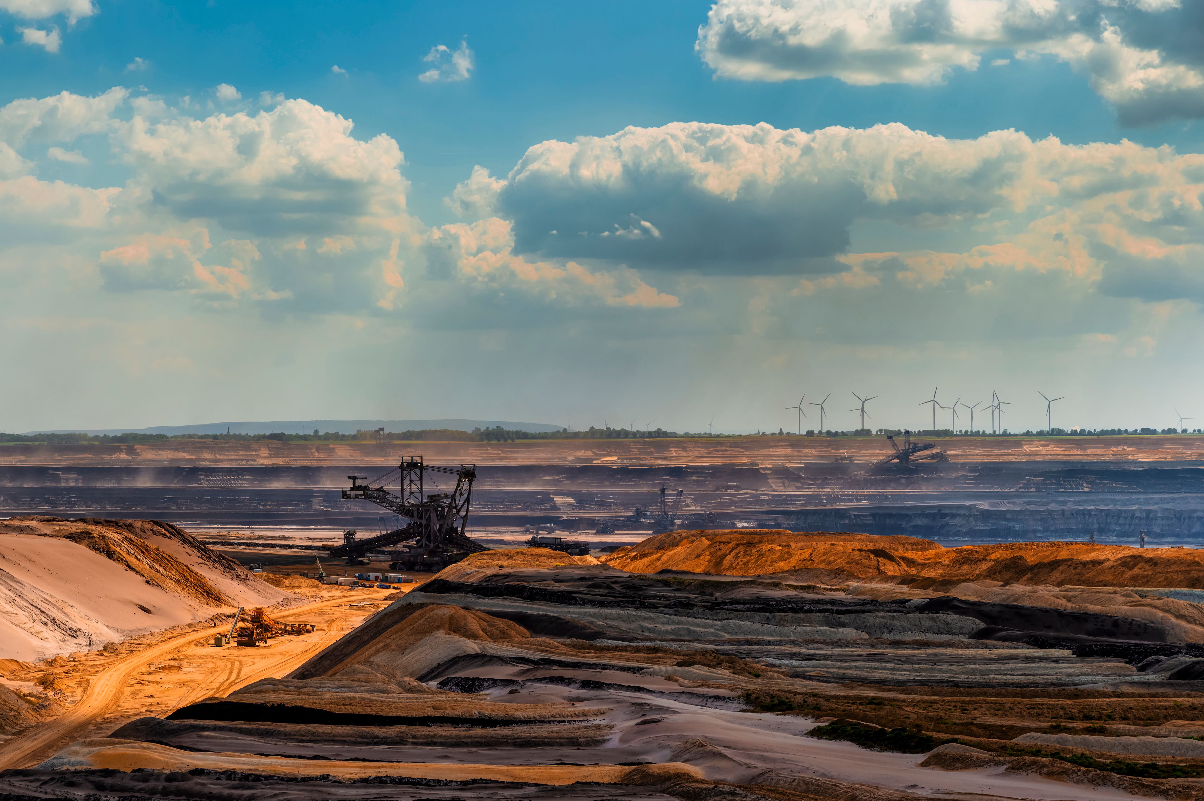 Mine à ciel ouvert de lignite avec excavatrices, sous un ciel nuageux, et éoliennes à l’horizon : le contraste entre extraction et énergie "verte".