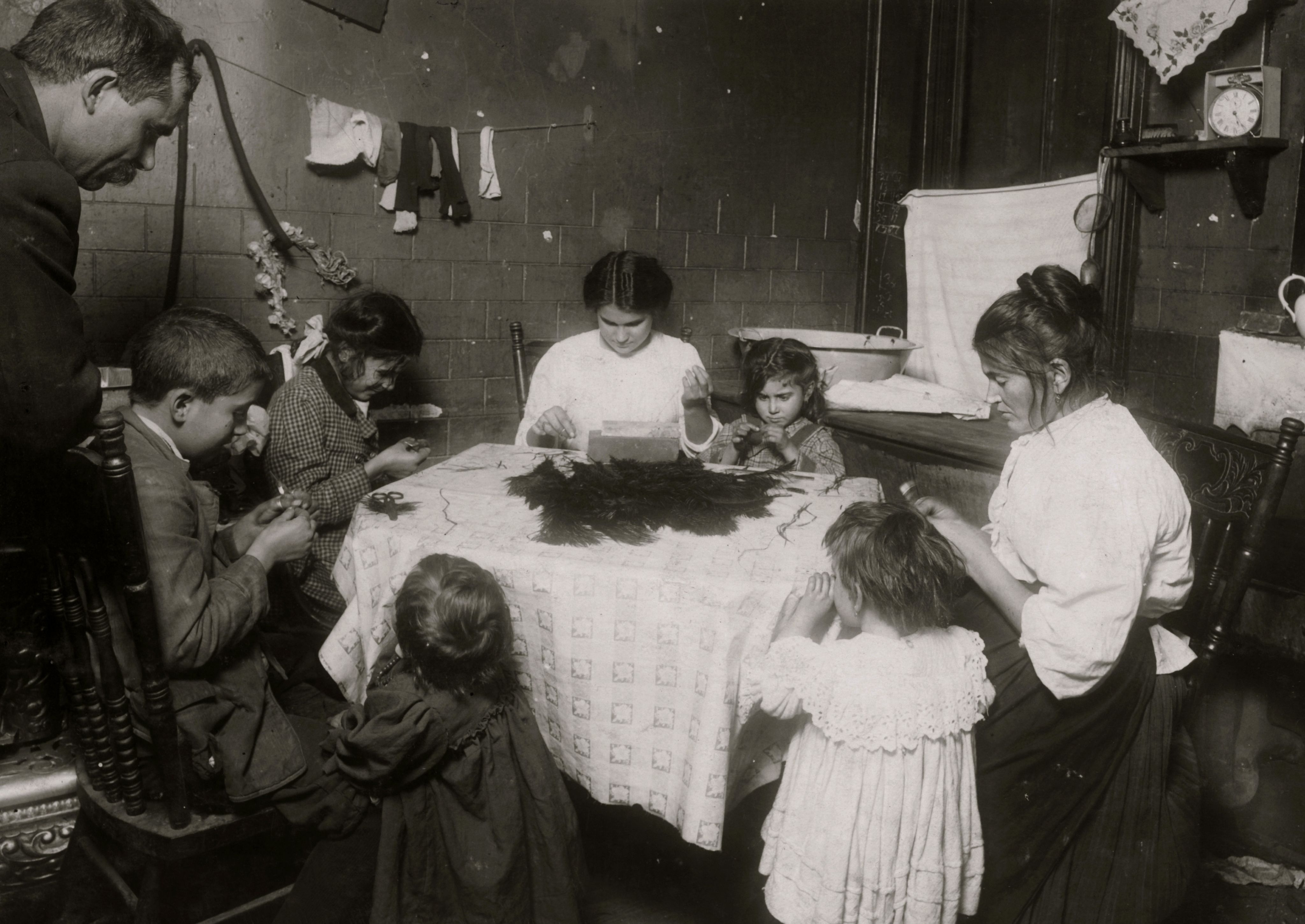 Sous le regard de leur père, six enfants nouent des plumes avec leur mère à une table dans une cuisine exiguë d'un immeuble, New York, New York, 1911.