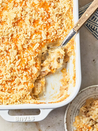 A close-up of a partially scooped dairy-free cheesy potato casserole in a white baking dish, showing a creamy and golden interior. A serving spoon rests in the dish, highlighting the creamy and cheesy texture under the crunchy chip topping.
