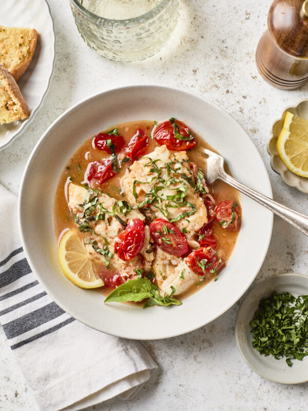 A bowl of cooked fish fillets topped with chopped herbs, cherry tomatoes, and a lemon slice, served with broth. Nearby are a glass of water, a plate with bread, a pepper grinder, lemon wedges, and a napkin.
