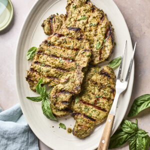 Grilled pork chops with visible grill marks on a white oval plate, garnished with fresh basil leaves. A meat fork rests beside the chops, and a light blue napkin is partially visible nearby.
