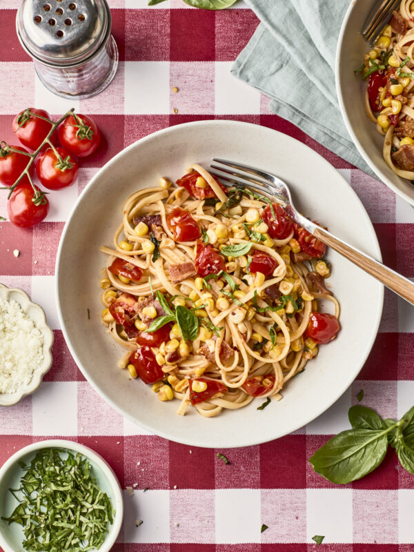 A bowl of pasta with cherry tomatoes, corn, basil, and cheese sits on a red-and-white checkered tablecloth, surrounded by fresh ingredients and a fork.