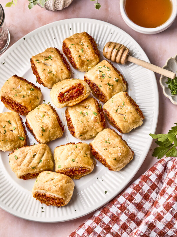 A white plate with several golden-brown sausage rolls, one cut open to show the filling. A honey dipper rests nearby, with a bowl of honey, green herbs, and a red-checked napkin beside the plate.