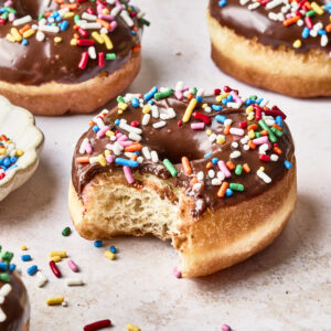 Chocolate-glazed donuts topped with colorful sprinkles sit on a light surface. One donut in the foreground has a bite taken out of it, with sprinkles scattered around.