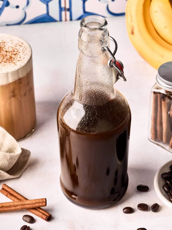 A glass bottle filled with dark liquid sits on a white surface, surrounded by coffee beans, cinnamon sticks, a jar with cinnamon sticks, a frothy iced coffee, and a bunch of bananas in the background.