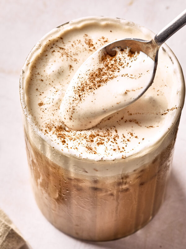 A glass of iced coffee topped with thick, creamy foam and a sprinkle of cinnamon, with a spoon scooping up some of the foam.