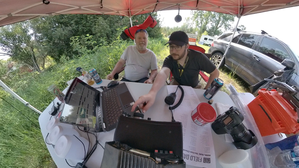 Two operators at a Field Day station under a red canopy with radios, laptop, and logging gear