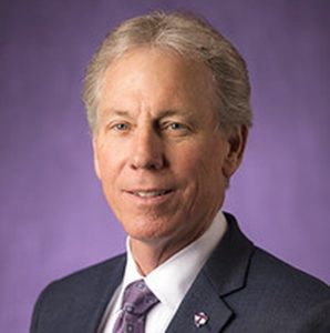 Portrait of a man with gray hair, wearing a suit, against a purple background.