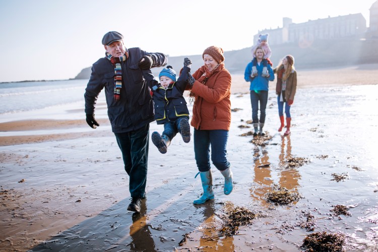 Multi-generation family walking along the beach with grandparents swinging a child.
