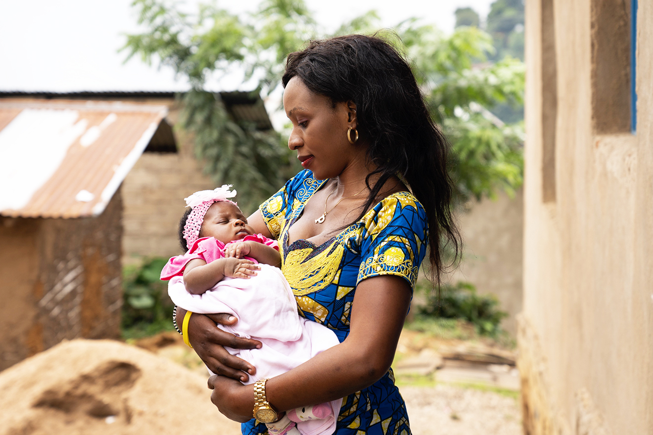 Ngitsi Kavugho was pregnant when she was infected with the Ebola virus. She and her baby survived. A young woman stands amidst homes, cradling her baby girl in her arms whilst gazing at her with love.