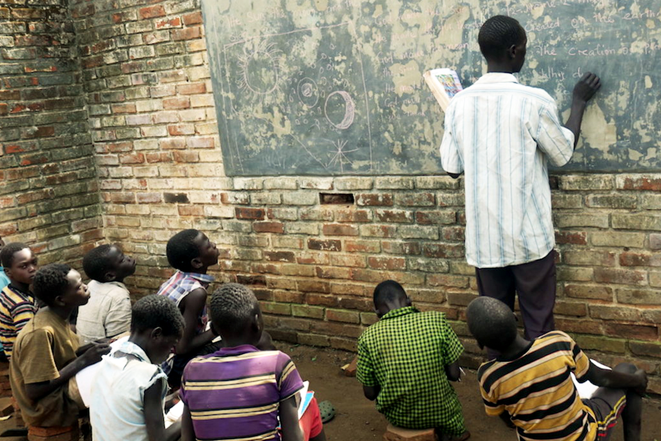 Previously displaced children have returned home to Gunyoro. Schooling has resumed, albeit under challenging circumstances. Classroom scene with teacher writing on a chalk-board whilst children sit on stools and look on.
