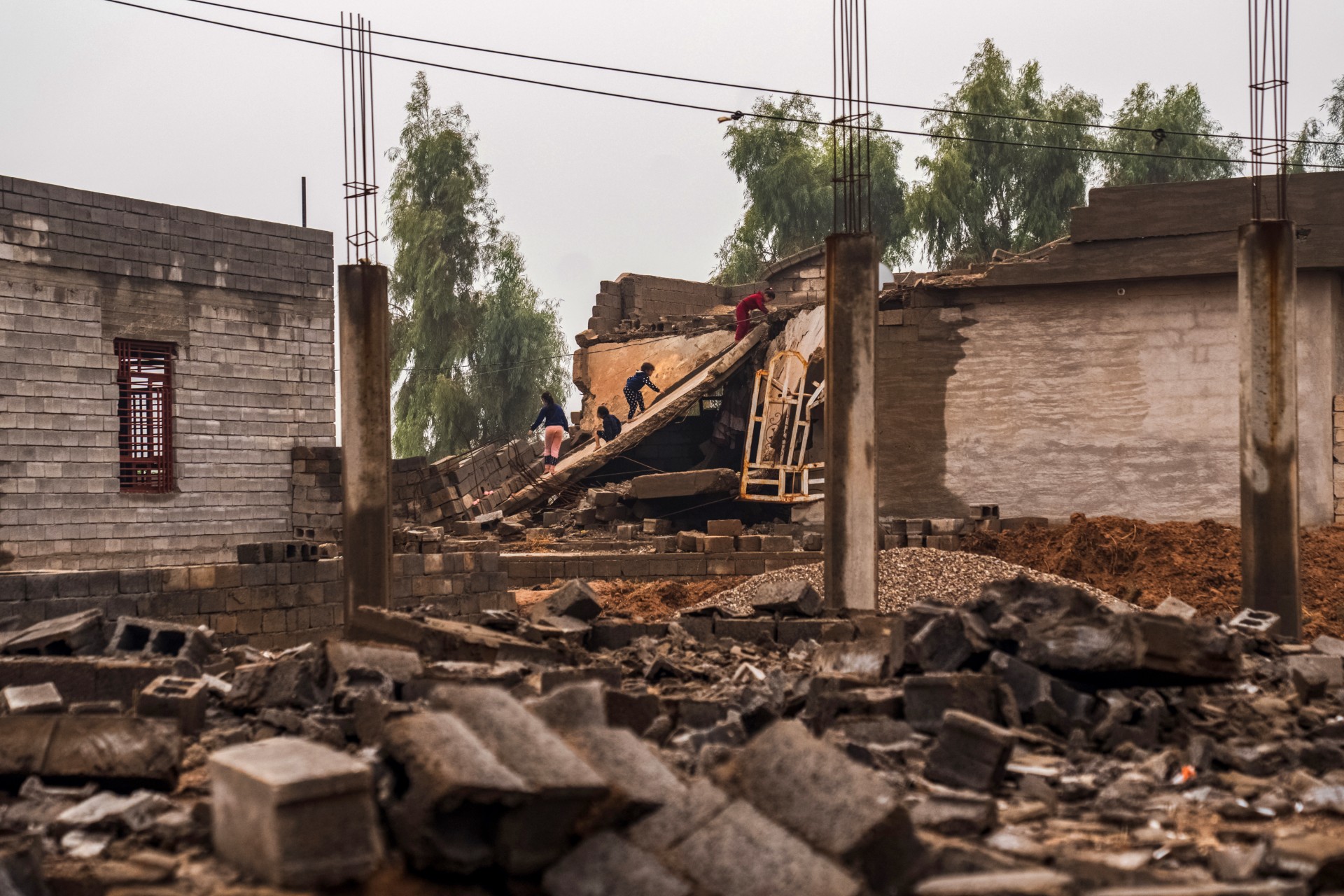 People climbing over debris between damaged buildings in a heavily ruined area.