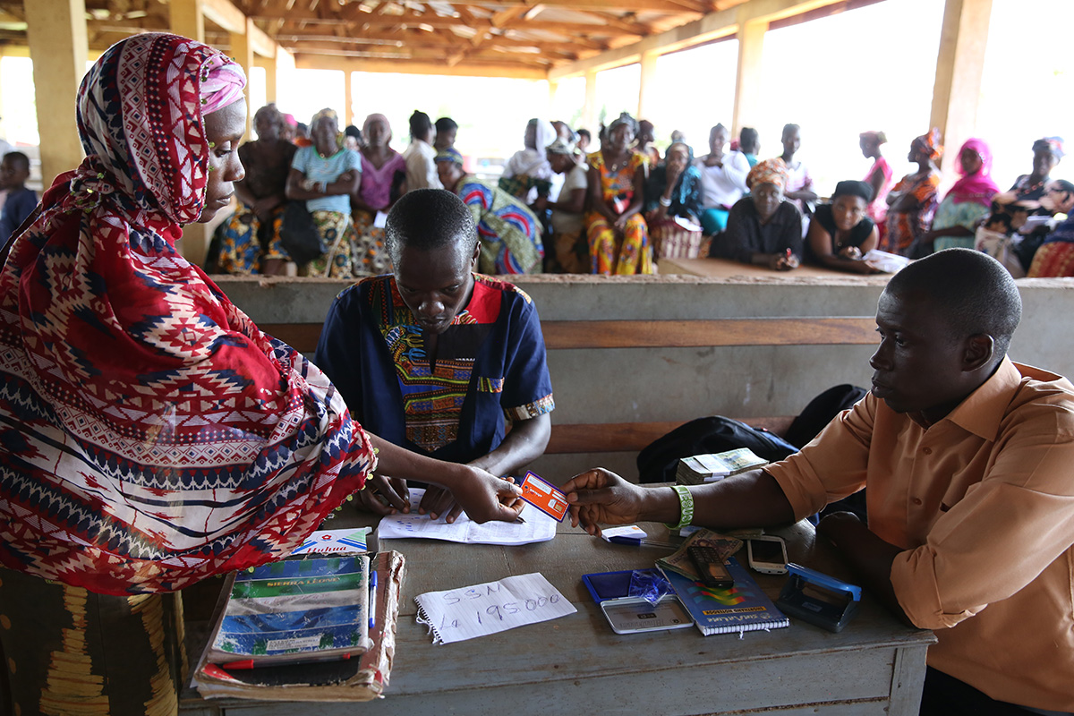 Receiving a cash transfer payment in Freetown, Sierra Leone. Photo © Dominic Chavez/World Bank Three people at a desk exchanging a card and making paper records.