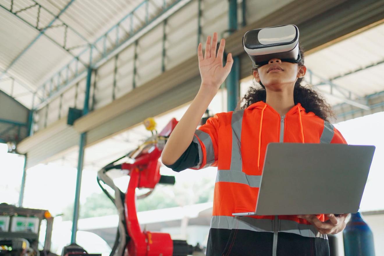 Mujer con un casco de realidad virtual, sosteniendo una computadora portátil en su mano izquierda y dibujando un espacio imaginario con su mano derecha.