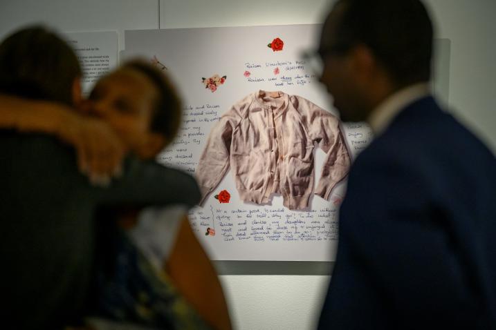 A view of the opening of the exhibit “Stories of Survival and Remembrance – A Call to Action for Genocide Prevention”. As part of an exhibit on genocide prevention, a framed shirt surrounded by text is shown in the background, while two people embracing in the foreground are blurred.