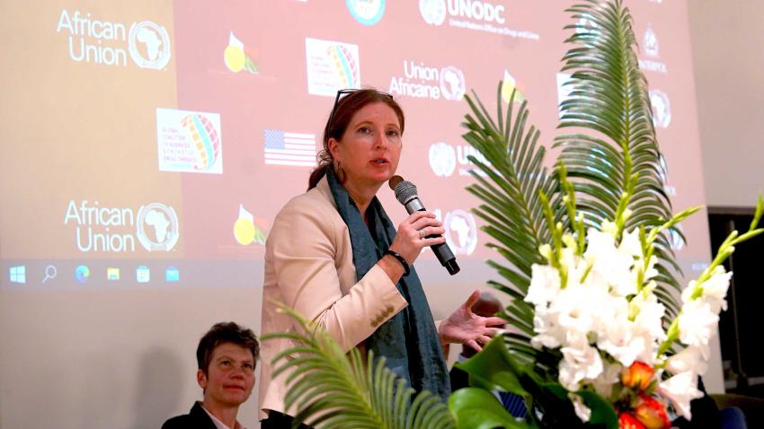 Carmen holds a microphone at a stage, and as she speaks, a woman is seen seated behind her intently listening.