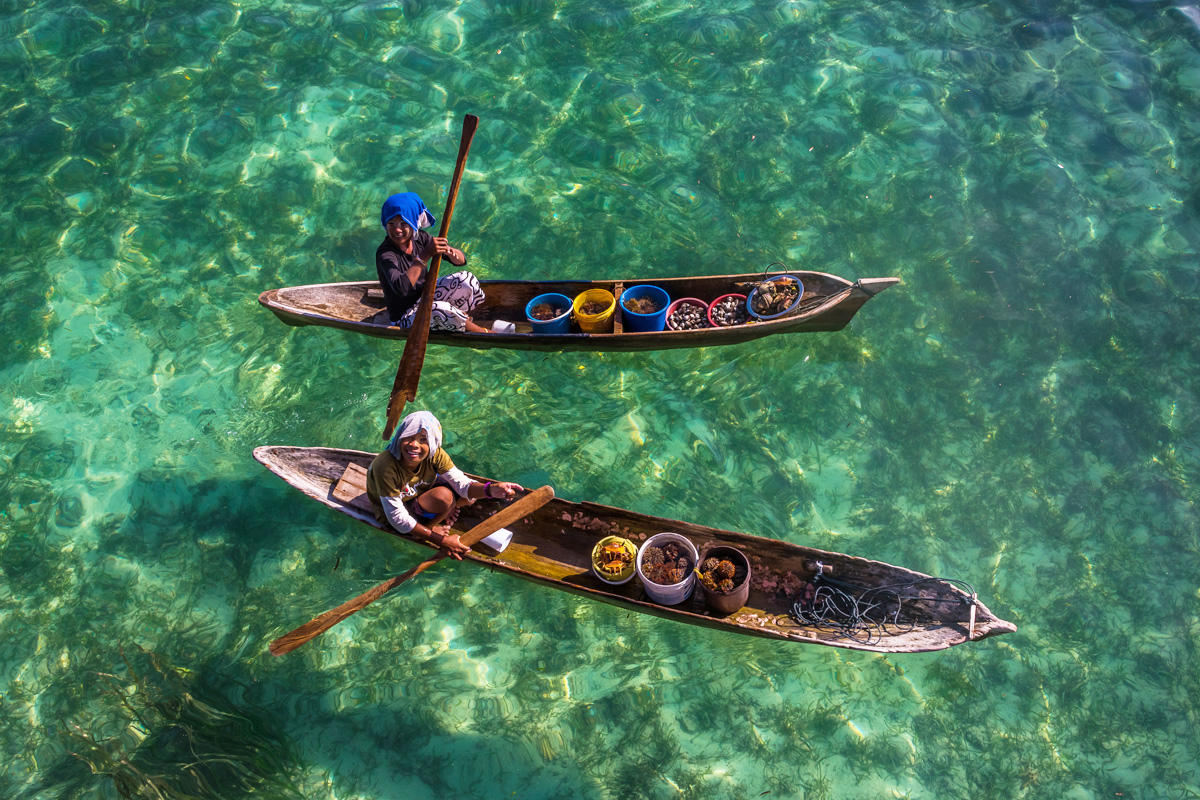 An overhead shot of emerald green waters with two small rowing boats each containing a young woman and their catch of the day