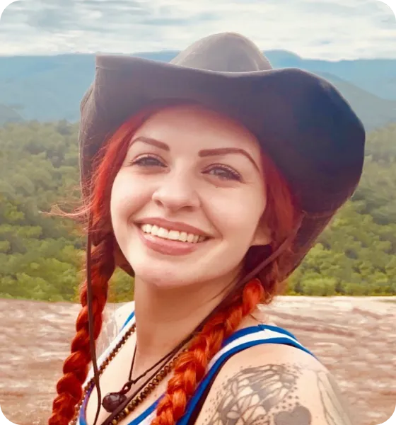 Smiling redhead in cowboy hat, braided hair, and necklace; mountain backdrop.